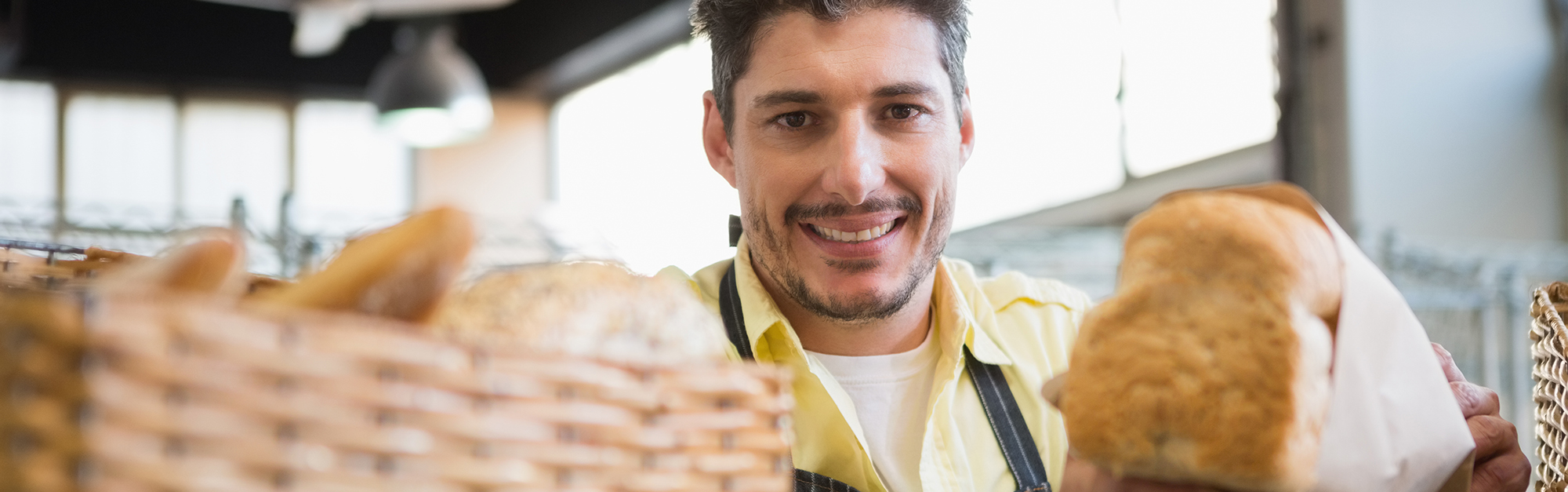 Ein lächelnder Mann über 25 mit dunklen Haaren und Dreitagebart steht hinter einer Bäckertheke und guckt durch Brötchen in die Kamera